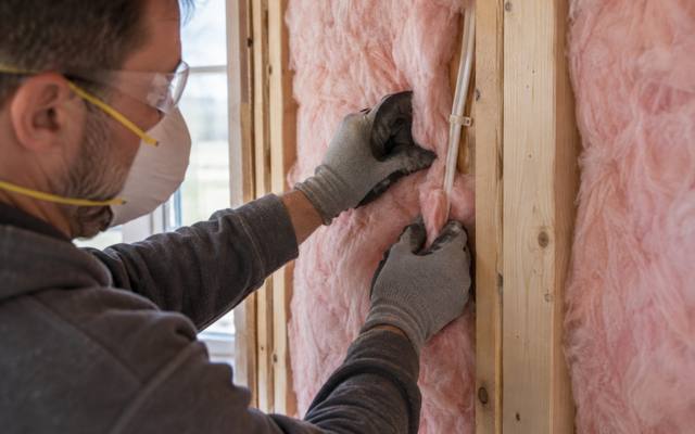 Worker in gloves and a dust mask fitting pink insulation batts between wall studs beside a window
