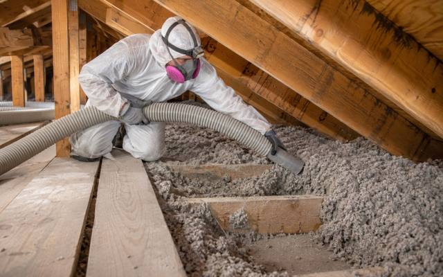 Worker in a white protective suit and respirator holding a large hose over grey loose-fill insulation between attic joists