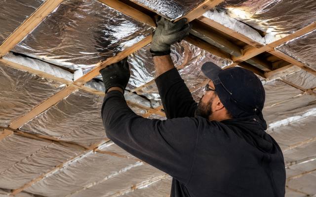 Worker in a black cap pressing foil-faced insulation into the framing of a sloped ceiling
