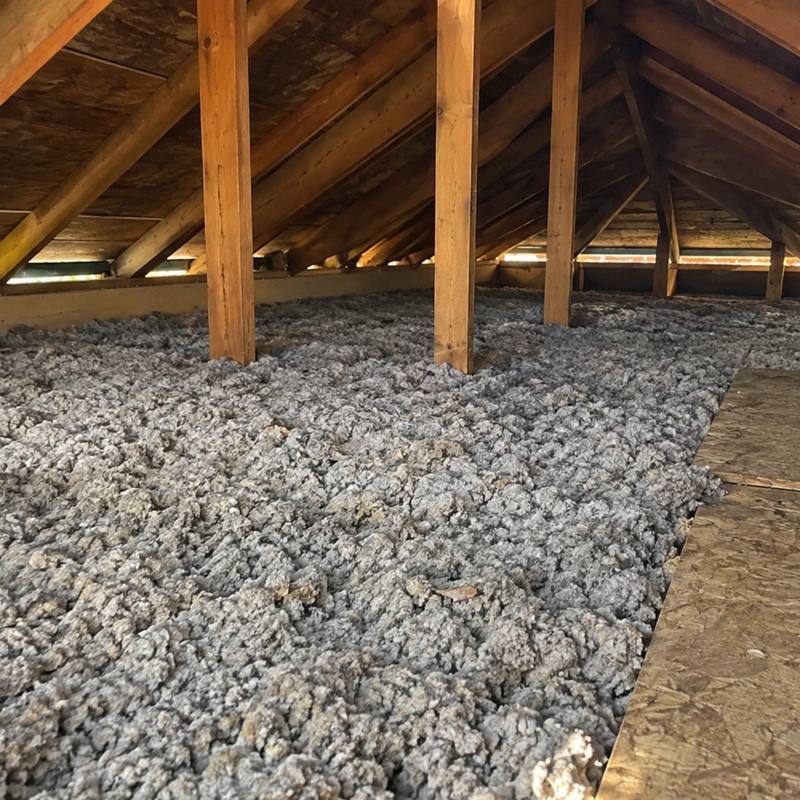 Grey loose-fill insulation spread deep across an attic floor with a strip of plywood laid over it and vents along the eave